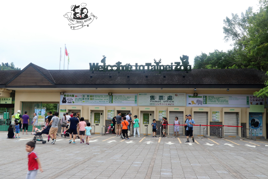 Wide angle view of Taipei Zoo entrance area with multiple ticket counter windows on the right side of the pathway and automated entry gates on the left side with visitors queuing